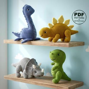 May include: Four crocheted dinosaur toys in blue, yellow, gray, and green are displayed on wooden shelves. The dinosaurs include a long-necked blue dinosaur, a yellow stegosaurus, a gray triceratops, and a green T-Rex. A PDF pattern is available.