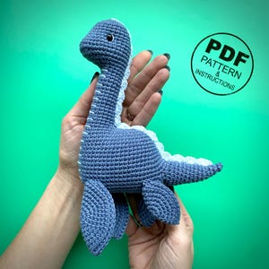 May include: A handmade blue crochet dinosaur toy with a light blue belly and accents. The toy has small black eyes and is held in a person's hands. A circular graphic with the text "PDF PATTERN & INSTRUCTIONS" is in the background.