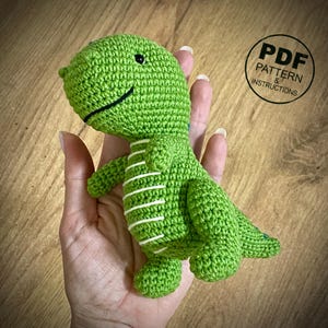 May include: A green crocheted dinosaur toy with white stripes on its belly. The dinosaur has a smiling face with black eyes and a black mouth. The image includes a circular graphic that says "PDF PATTERN & INSTRUCTIONS".