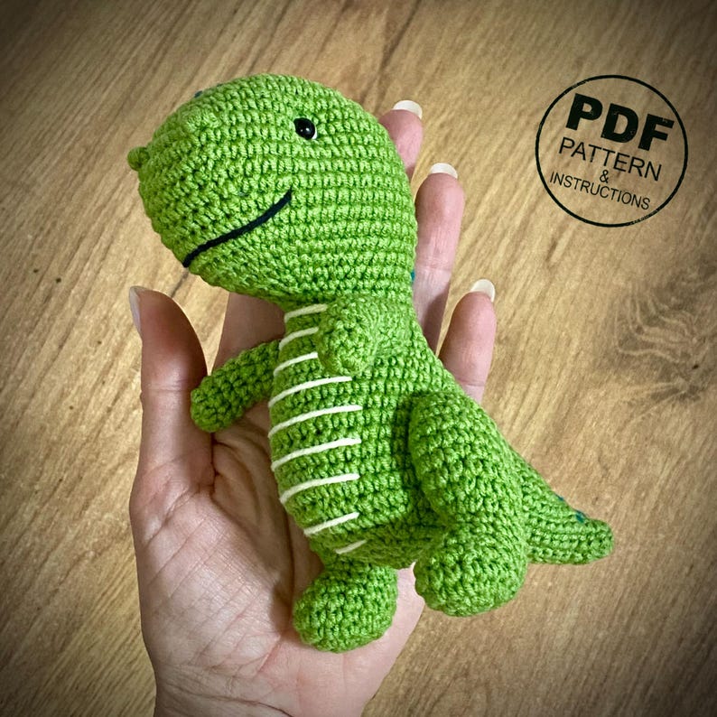 May include: A green crocheted dinosaur toy with white stripes on its belly. The dinosaur has a smiling face with black eyes and a black mouth. The image includes a circular graphic that says "PDF PATTERN & INSTRUCTIONS".
