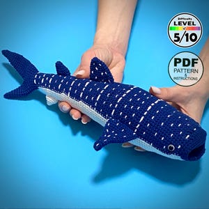 May include: A handmade, blue and white crocheted whale shark toy with white spots. The toy is held against a blue background. The image includes a Difficulty Level 5/10 graphic and a PDF Pattern & Instructions graphic.