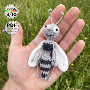 May include: A small, handmade crochet insect toy featuring a grey and black striped body, white wings, and two antennae. The head has two round white eyes with black pupils and a stitched smile. Text on the image reads Difficulty LEVEL 4/10 and PDF PATTERN & INSTRUCTIONS.