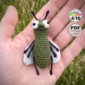 Puede incluir: Un pequeño juguete de insecto tejido a ganchillo en color verde oliva con ojos blancos, pupilas negras, antenas negras y alas gris claro. La imagen incluye el texto Difficulty Level 4/10 y PDF Pattern & Instructions.