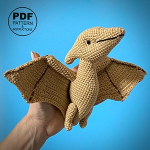 May include: A tan crocheted Pterodactyl toy with outstretched wings, black eyes, and a detailed beak. The wings have a darker brown trim. The image includes a circular graphic with the text "PDF PATTERN & INSTRUCTIONS".