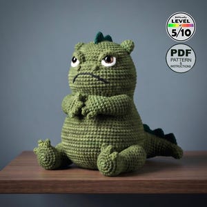 May include: A green crocheted monster doll with a grumpy expression, brown eyes, and a dark green dorsal fin. The image includes a circular badge stating Difficulty Level 5/10 and a second badge indicating PDF Pattern & Instructions for this item.