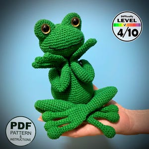 May include: A handmade green crochet frog with large, golden eyes and outstretched arms. The frog is sitting with its legs extended. The image includes a difficulty level of 4/10 and a PDF pattern and instructions label.