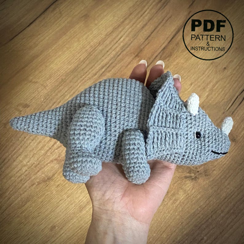 May include: A crocheted triceratops dinosaur in light gray, held in a hand. The dinosaur has three white horns, black eyes, and a smiling mouth. The image includes a circular graphic that says "PDF PATTERN & INSTRUCTIONS."