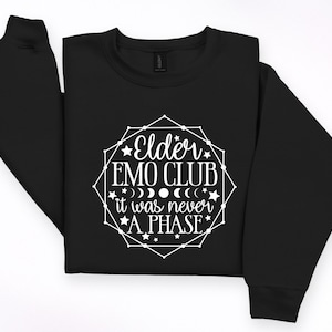 May include: Black sweatshirt with the text "Elder EMO CLUB it was never A PHASE" in a white geometric design. The design includes stars, moons, and the text in a stylized font. The sweatshirt is folded on a white surface.