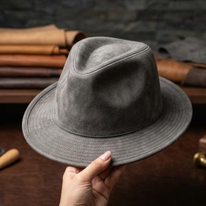 Peut inclure: Un chapeau fedora gris avec une couronne structurée et un large bord. Le chapeau est présenté devant un fond de cuirs et d'outils, suggérant un produit artisanal. La couleur du chapeau est un gris mat, et il semble être fait d'une matière douce.