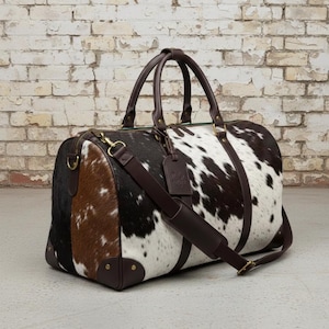 May include: A cowhide duffel bag with brown leather detailing. The bag has panels of black, brown, and white cowhide, a brown leather shoulder strap, and two brown leather handles. It features a zip closure and a leather tag.