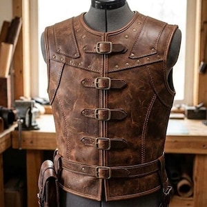 May include: A brown leather vest featuring a round neckline, structured shoulder panels, and a front closure with four adjustable metal buckles. The garment includes a horizontal waist belt with a matching buckle and a small attached leather pouch.