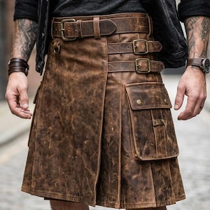 May include: A brown leather kilt featuring a distressed finish, a wide waistband with a buckle, two side straps with metal buckles, and a large cargo pocket on the front right side. The garment includes vertical pleats and visible stitching.