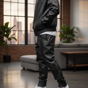 Pode incluir: Uma pessoa vestindo uma jaqueta de couro preta e cal&ccedil;as combinando. A jaqueta tem bot&otilde;es e as cal&ccedil;as t&ecirc;m cintura com cord&atilde;o. T&ecirc;nis brancos completam o look. O conjunto est&aacute; em um interior moderno.