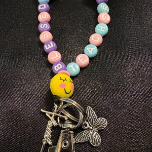 May include: A beaded lanyard with the words "BLESSED TEACHER" spelled out in colorful round beads. The lanyard features a yellow smiley face bead, a silver-tone clasp, and a butterfly charm. The lanyard is designed for teachers.