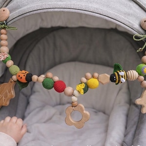 Peut inclure: Un jouet de poussette en bois pour bébé avec divers éléments en crochet et en bois colorés. Le jouet présente des motifs de coccinelle, d'abeille et de fleur, ainsi que des perles en bois et des formes de feuilles et de papillons. Le jouet est fixé à une poussette grise.