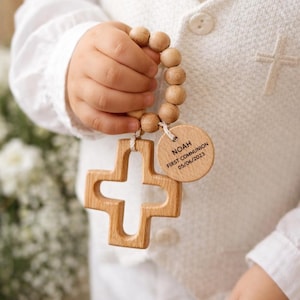 Op de afbeelding: Een kind houdt een houten kruis en rozenkrans kralen vast. Het kruis is lichtbruin met een uitgesneden middenstuk. Een klein houten label leest "NOAH FIRST COMMUNION 05/06/2023". Het kind draagt een witte outfit.