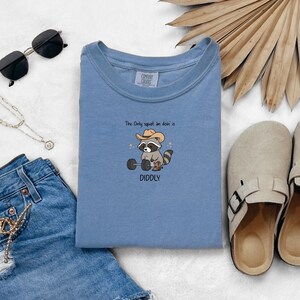 Puede incluir: Sudadera azul claro con el texto "The Only squat im doin' is DIDDLY" y un mapache de dibujos animados con sombrero de vaquero levantando pesas. La sudadera está rodeada de accesorios, incluyendo gafas de sol y un collar.