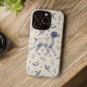 May include: A phone case with a cream background and a blue hunting dog design. The case features a running dog, birds, and hunting horns. The phone case is on a wooden surface.