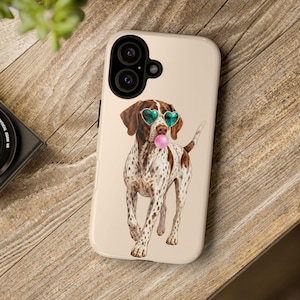 Puede incluir: Una funda de smartphone beige con una ilustración impresa de un perro manchado marrón y blanco que lleva gafas de sol en forma de corazón de color verde azulado y sopla una burbuja de chicle rosa. Tiene un recorte negro para la cámara.