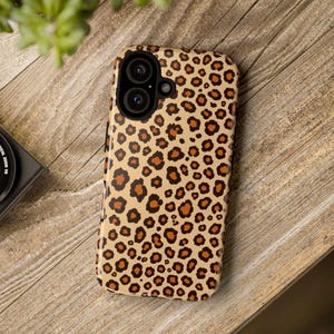 May include: A tan phone case with a leopard print design. The case features a pattern of brown and dark brown spots. The phone case is on a wooden surface with a camera and plant in the background.