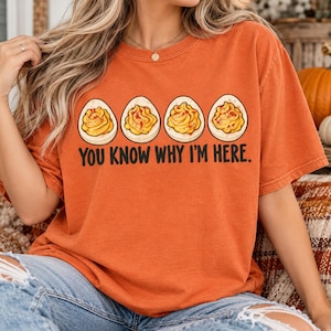 May include: An orange short-sleeve t-shirt featuring a graphic of four halved deviled eggs in a row. Below the eggs, the black text reads, "YOU KNOW WHY I'M HERE." The shirt has a crew neckline and a relaxed fit, showcasing a solid color design.