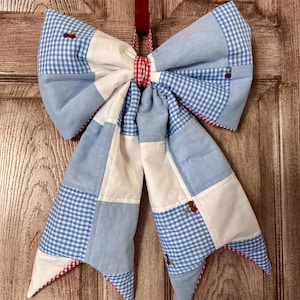 May include: A large, decorative bow made of patchwork fabric in shades of blue and white gingham, with red accents. The bow features embroidered cherries and is designed to hang. The bow is attached to a red ribbon.