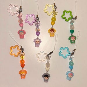 May include: A collection of colorful phone charms. Each charm features a star-shaped top, a string of beads, and a small figurine in a hooded outfit. The charms come in various colors, including pink, purple, yellow, green, orange, clear, and blue.