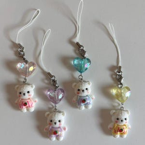 Op de afbeelding: Vier witte teddybeer bedels met hartvormige hangers. Elke beer draagt een verschillend gekleurde outfit met een aardbeienontwerp. De hangers zijn in roze, blauwe en gele tinten, bevestigd aan een zilveren sluiting en een wit koord.