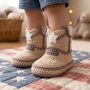 May include: A pair of beige crochet booties designed to resemble cowboy boots. Each boot features a white crocheted star appliqué on the side, brown yarn detailing along the seams, and a rounded toe. The booties are worn on feet over a quilt.