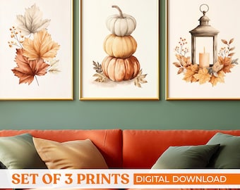 May include: Set of three framed prints with autumn themes. One print features a stack of pumpkins, another shows a lantern with a candle, and the third displays fall leaves. The prints have gold frames and are displayed above a red sofa.