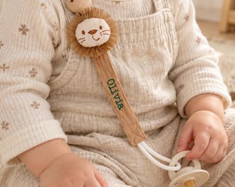 May include: A beige fabric pacifier clip featuring a plush lion head with a brown fringe mane and a wooden bead attachment. The strap is embroidered with the name Olivia in green thread and connects to a cream-colored silicone pacifier.