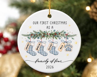 May include: A round white ceramic Christmas ornament featuring the text "OUR FIRST CHRISTMAS AS A family of four 2026". The design includes green pine branches with red berries, three blue plaid stockings, and a paw print tag labeled James, Amelia, Olivia, and Milo.