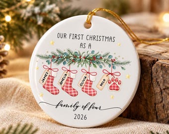May include: A round white ceramic Christmas ornament featuring the text "OUR FIRST CHRISTMAS AS A family of four 2026". The design includes green pine branches with red berries, four red plaid stockings with name tags, and a red plaid paw print.