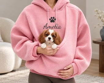 May include: A light pink, long-sleeved, hooded sweatshirt featuring a large front pouch pocket. The chest area displays a black paw print graphic above the name Amelia in a cursive font. A small brown and white puppy sits inside the pouch.