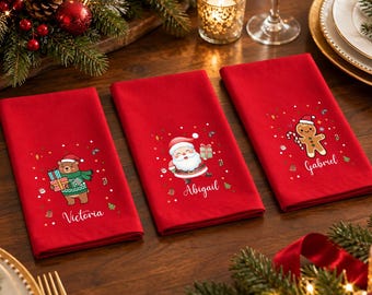 May include: Three rectangular red cloth napkins featuring festive holiday graphics. One napkin shows a bear with gifts and the name Victoria, one shows Santa Claus and the name Abigail, and one shows a gingerbread man and the name Gabriel.