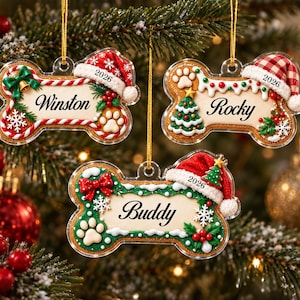 May include: Three bone-shaped Christmas ornaments made of clear acrylic. Each ornament features a festive design with a Santa hat, holly, and a paw print. The names Winston, Rocky, and Buddy are printed in black script, with the year 2026 included.