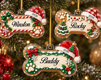 May include: Three bone-shaped Christmas ornaments made of clear acrylic. Each ornament features a festive design with a Santa hat, holly, and a paw print. The names Winston, Rocky, and Buddy are printed in black script, with the year 2026 included.