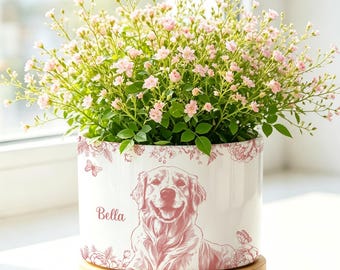 May include: A white cylindrical ceramic planter featuring a pink line-art illustration of a dog, the name Bella, and floral accents. The planter sits on a circular wooden base and contains a small rose bush with numerous light pink flowers.