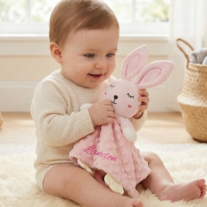 Könnte beinhalten: Ein Baby hält eine rosa Kuscheldecke mit einem Hasenmotiv, auf der der Name "Landon" gestickt ist. Der Hase hat lange Ohren und ein süßes Gesicht. Das Baby trägt einen cremefarbenen Strampler und sitzt auf einem flauschigen weißen Teppich.