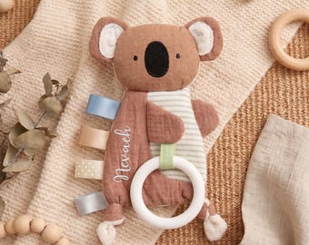 May include: A brown koala-shaped security blanket featuring a white striped belly, a black embroidered nose, and the name Nevaeh stitched in white. The toy includes four colorful fabric ribbon tags on the side and a white circular teething ring.