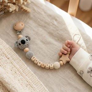 May include: A wooden baby pacifier clip with a gray crocheted koala bear, wooden beads, and the name "Zavier" spelled out. The clip is attached to a star-shaped wooden teether held by a baby.