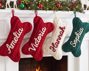 May include: Four bone-shaped Christmas stockings made of cable-knit fabric. Two are red with white embroidered names Amelia and Victoria, one is cream with a red embroidered name Hannah, and one is dark green with a white embroidered name Sophia.