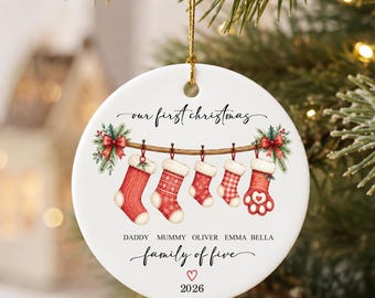 May include: A round white ceramic Christmas ornament featuring a printed illustration of five hanging stockings on a branch. The text reads our first Christmas, DADDY MUMMY OLIVER EMMA BELLA, family of five, and 2026. It includes a gold hanging cord.