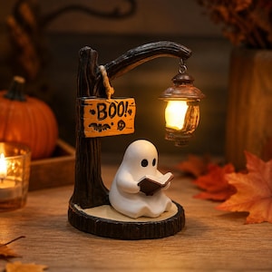 May include: A white ghost figurine sits on a dark brown base, reading a small book. A dark tree-like post holds a hanging lantern with a warm glowing light and a rectangular wooden sign with the text BOO! and small bat and skull illustrations.