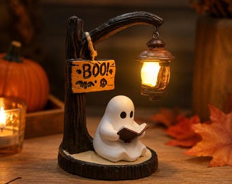 May include: A white ghost figurine sits on a dark brown base, reading a small book. A dark tree-like post holds a hanging lantern with a warm glowing light and a rectangular wooden sign with the text BOO! and small bat and skull illustrations.