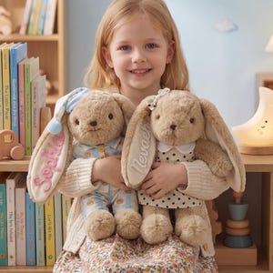 Puede incluir: Una niña sostiene dos conejos de peluche. Uno lleva un pijama azul y blanco y un gorro de dormir, el otro un vestido de lunares. Las orejas de los conejos están bordadas con nombres. La niña sonríe.