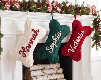 May include: Three bone-shaped Christmas stockings in cream, dark green, and red knit fabric. Each stocking features a cable knit pattern and a hanging loop. The cream stocking is embroidered with Hannah, the green with Sophia, and the red with Victoria.