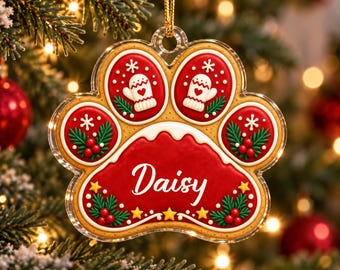 May include: A paw-shaped Christmas ornament with a clear acrylic border. The center features a red gingerbread-style design with the name Daisy in white script. It is decorated with white mittens, green pine sprigs, red berries, and yellow stars.
