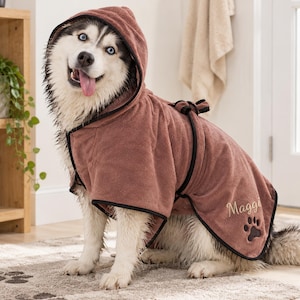 May include: A dusty rose dog bathrobe with a hood and black trim. The robe features the name Maggi embroidered in cursive script above a black paw print graphic on the side. The garment is secured with a matching tie belt around the waist.