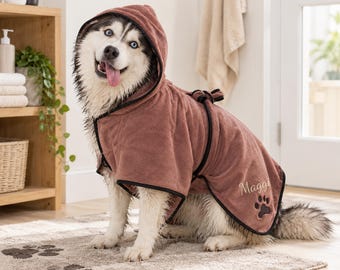 May include: A dusty rose dog bathrobe with a hood and black trim. The robe features the name Maggi embroidered in cursive script above a black paw print graphic on the side. The garment is secured with a matching tie belt around the waist.
